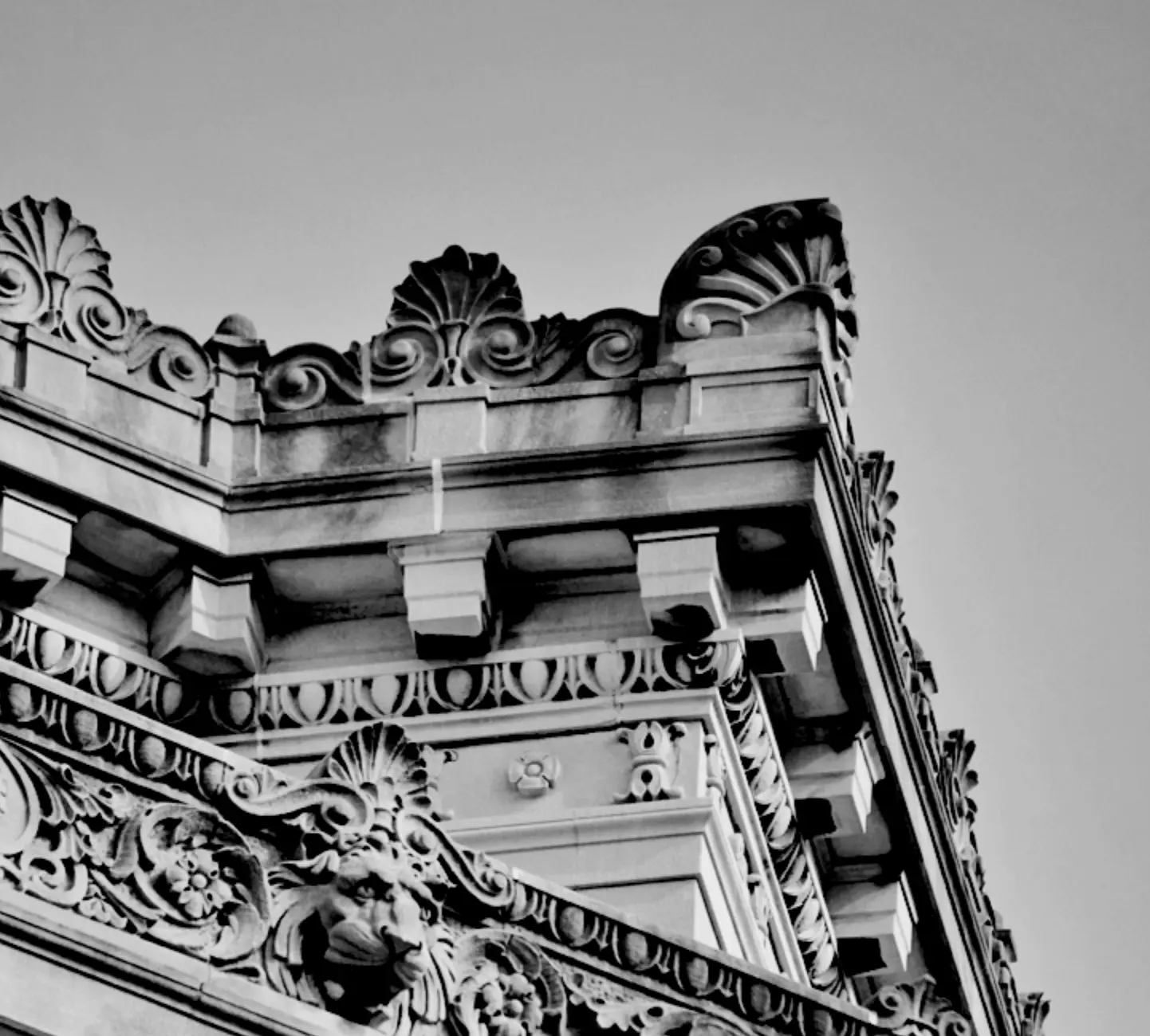 Black and white photo of an ornate building corner, showcasing detailed carvings of faces, floral patterns, and decorative stonework against a clear sky.