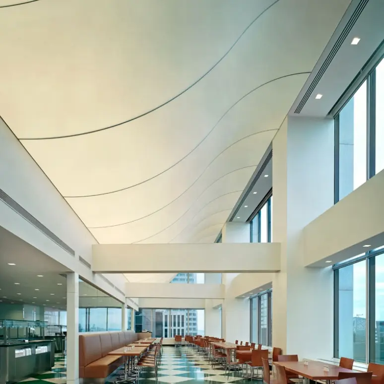 A modern cafeteria with large windows, high wavy ceilings, wooden chairs, and tables arranged in rows. Natural light fills the space, and the floor has a green and white checkered pattern.
