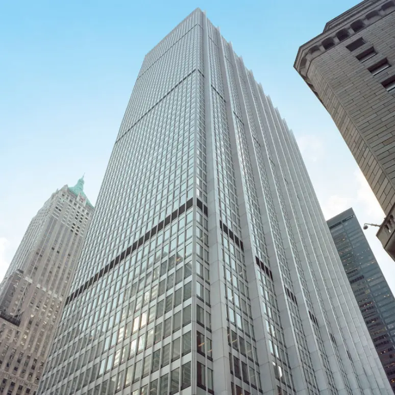 A tall, modern skyscraper with a grid of windows rises into a blue sky, flanked by other high-rise buildings with different architectural styles.