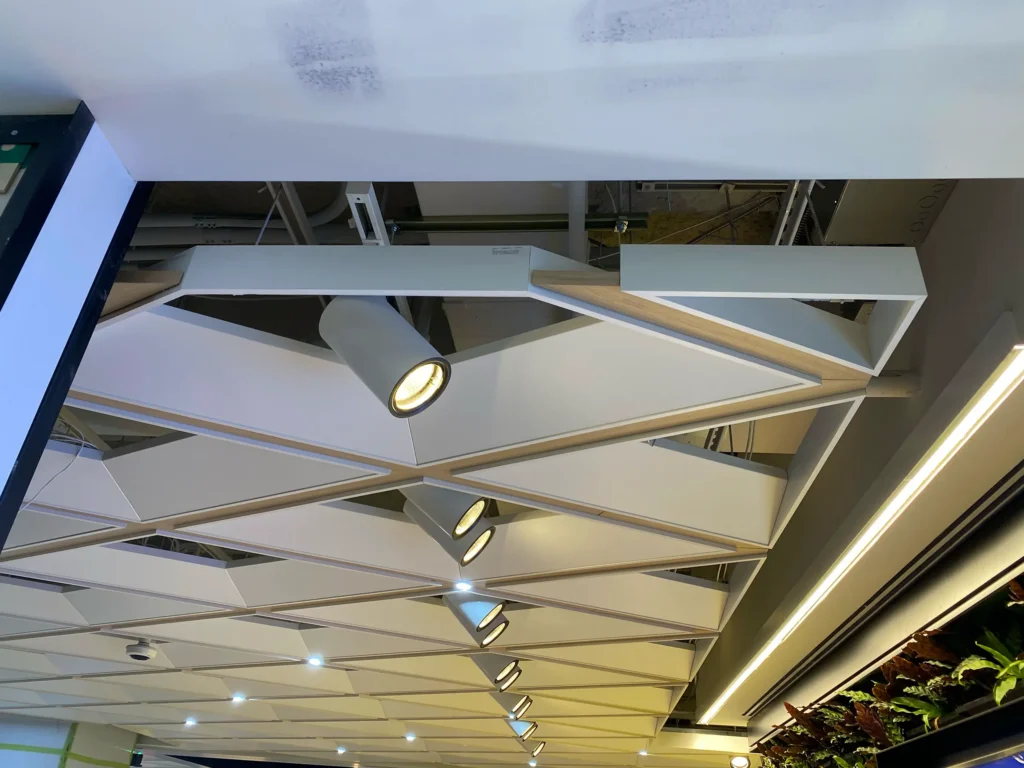 Ceiling with geometric white panels, cylindrical spotlights, exposed ducts above, and a section of greenery along the right edge. One spotlight is illuminated.