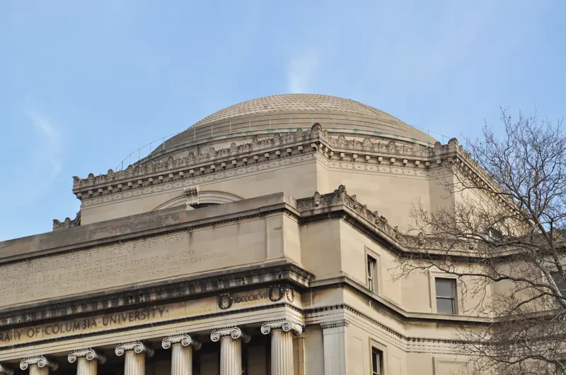 A neoclassical building with a large domed roof, engraved stone facade, and decorative columns, set against a clear blue sky and partially framed by bare tree branches.