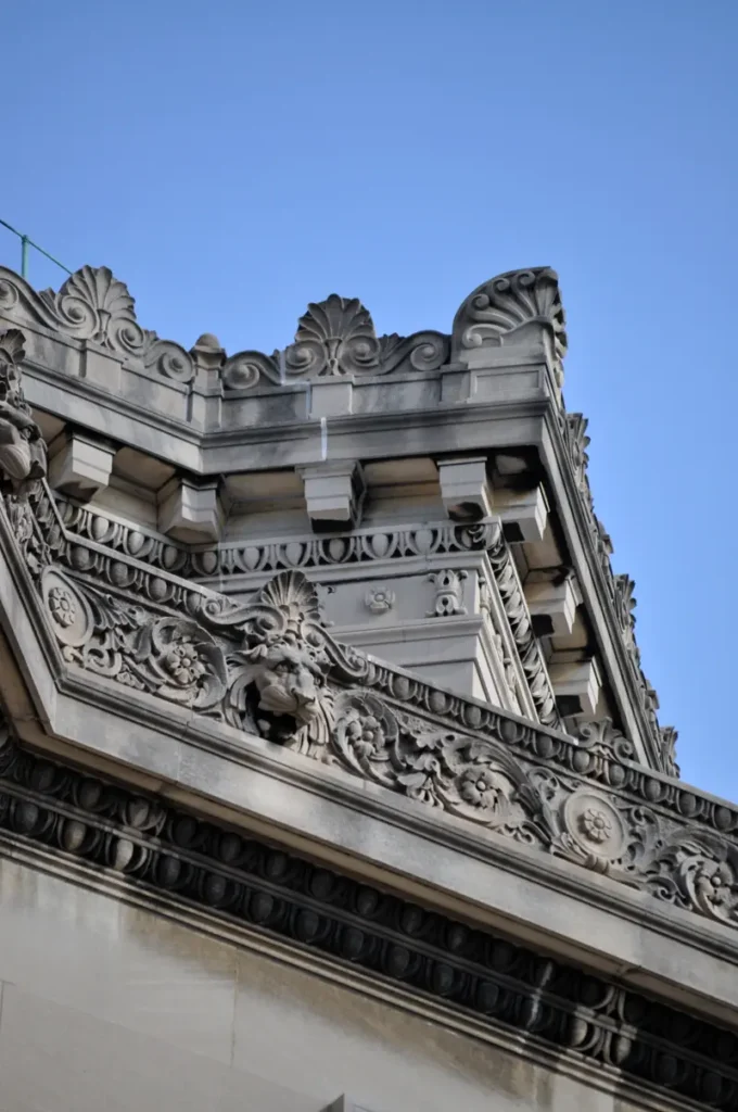The image shows an ornate stone building facade with intricate carvings of floral and scroll patterns along the roofline, set against a clear blue sky.