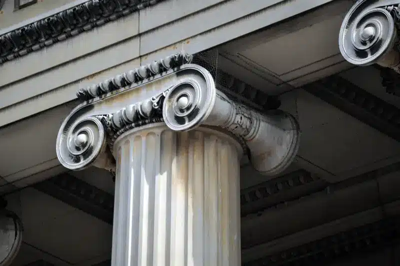 Close-up of a classical Ionic column with scroll-shaped volutes and decorative detailing, supporting the entablature of a building. The column is fluted and shows signs of weathering.