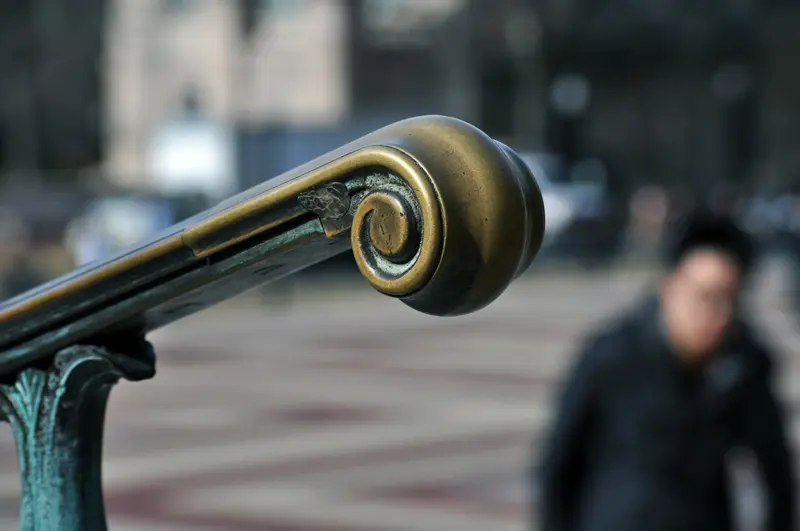 A close-up of a decorative bronze railing with a spiral design, set outdoors, with a blurred person and urban background in the distance.