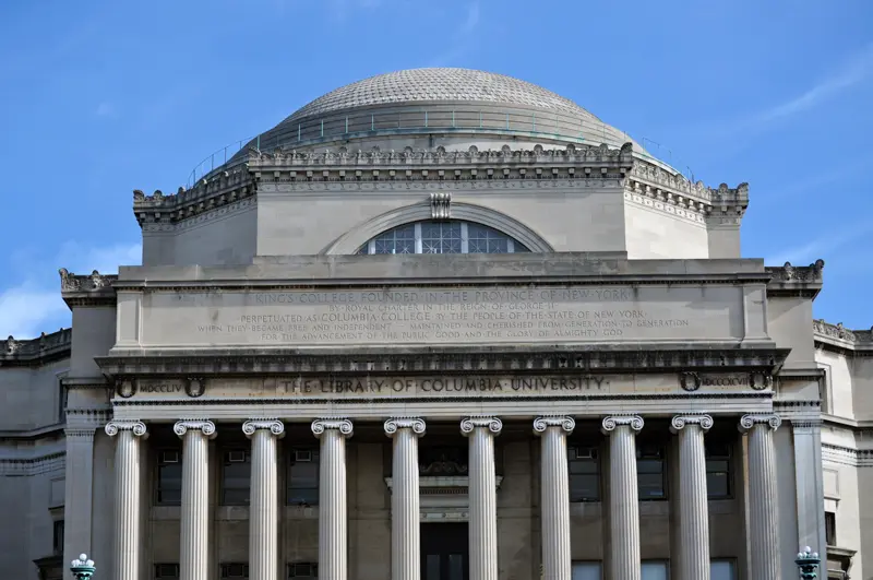 The photo shows the front of Columbia University's Butler Library, featuring grand columns, stone carvings, and a large dome under a blue sky. The building's name is engraved above the entrance.