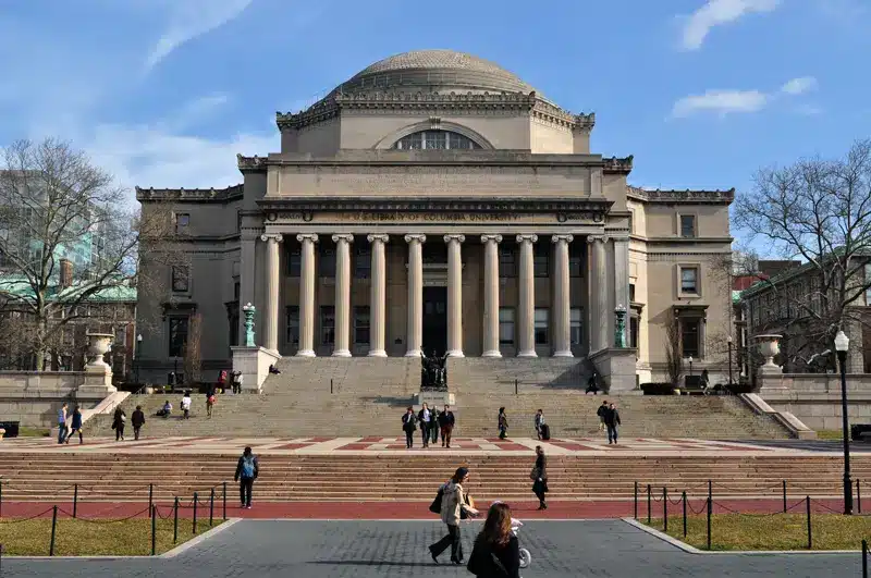 A large neoclassical building with tall columns and a domed roof, surrounded by steps and people walking in front of it on a clear day.