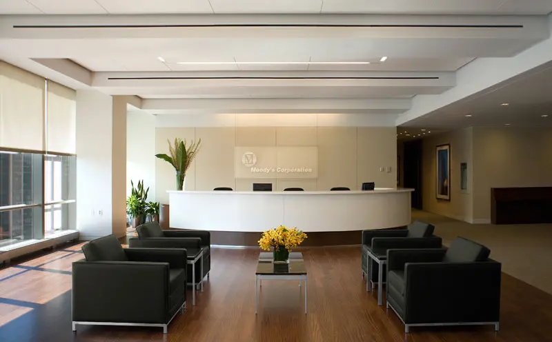 Modern office reception area with a white curved desk, four black chairs around a low table with yellow flowers, large windows with blinds, wood flooring, and minimalist décor.