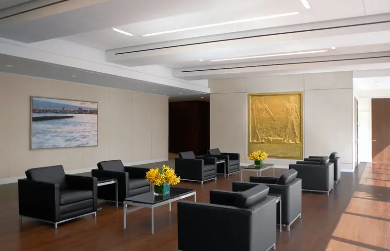 A modern waiting area with black armchairs, small glass tables with yellow flower arrangements, a large gold wall sculpture, and a landscape painting on light-colored walls and wooden flooring.