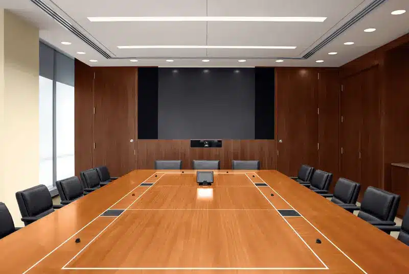 A modern, empty conference room with a large wooden table, black chairs around it, wood-paneled walls, a big screen at the front, and recessed ceiling lights.