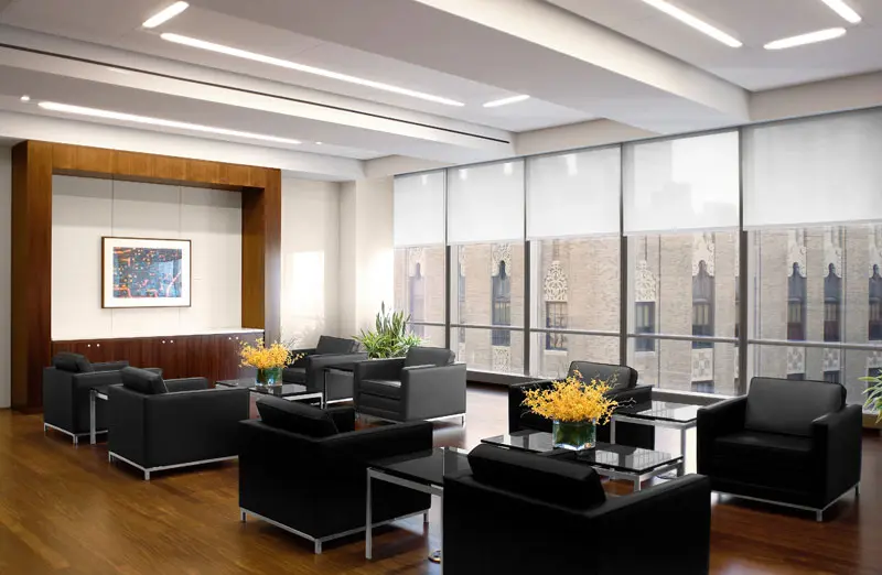 A modern office lounge with black armchairs, glass coffee tables, yellow flower arrangements, large windows with city views, wood flooring, and a framed artwork on the wall.