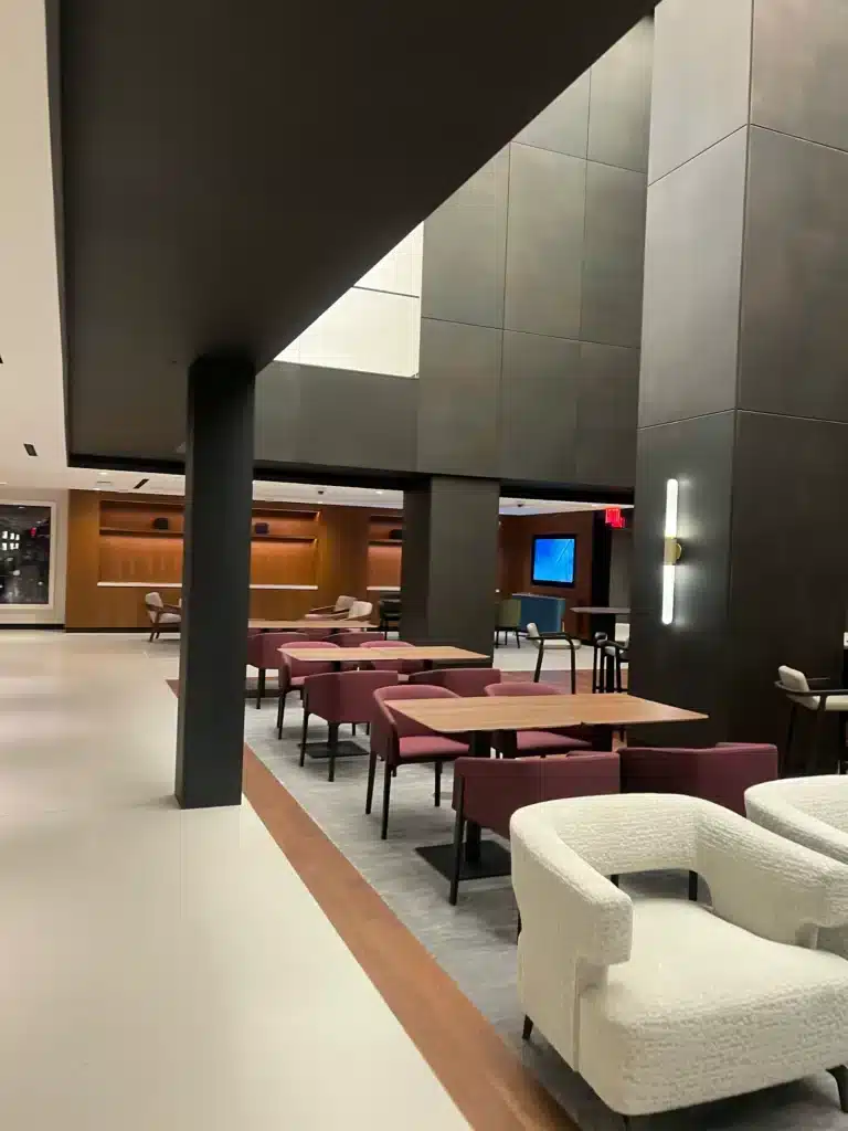 A modern, spacious lounge area with maroon and white chairs, wooden tables, soft lighting, high ceilings, and large dark wall panels; a TV is mounted on the far wall.