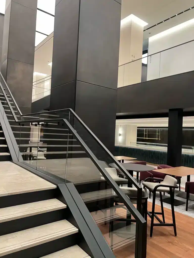 Modern office interior with a staircase featuring glass railings, dark walls, high ceilings, and an open seating area with tables, chairs, and a bar counter on the lower level.