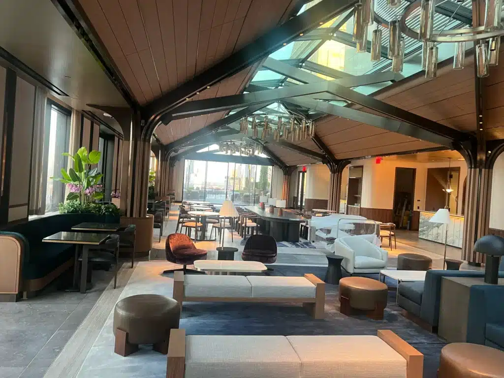 A modern, spacious lounge with high wooden ceilings, large windows, various seating areas including sofas, chairs, and tables, soft lighting, plants, and contemporary decor.
