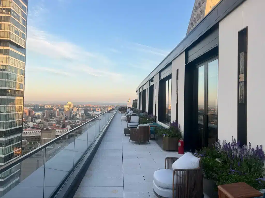 A rooftop terrace with modern outdoor seating, potted plants, and glass railing overlooks a cityscape at sunset, with tall buildings visible in the distance under a clear sky.