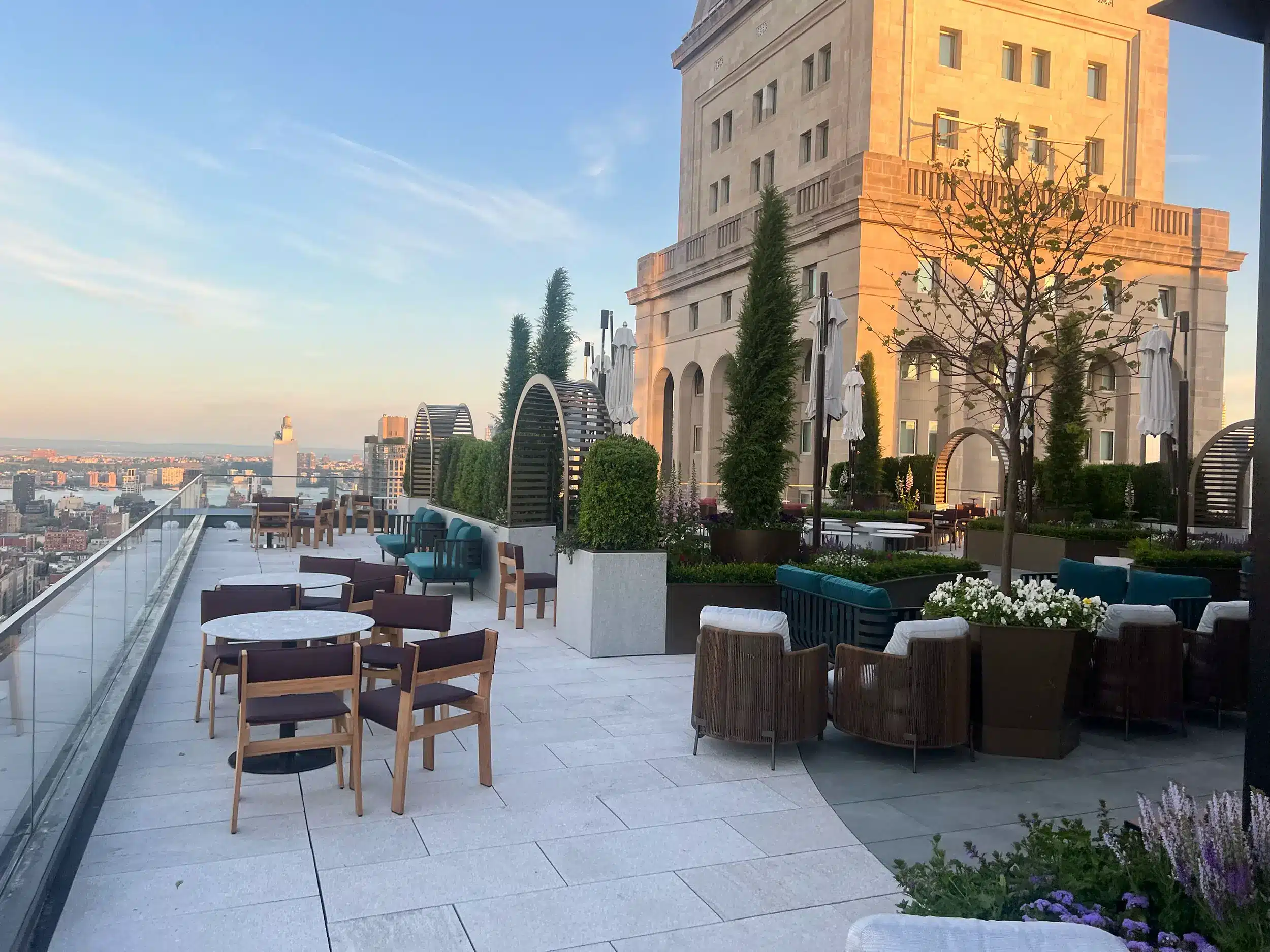 A stylish rooftop terrace with tables, chairs, and greenery offers city views at sunset. Modern outdoor furniture and tall potted plants create a relaxing, elegant atmosphere alongside a historic stone building.