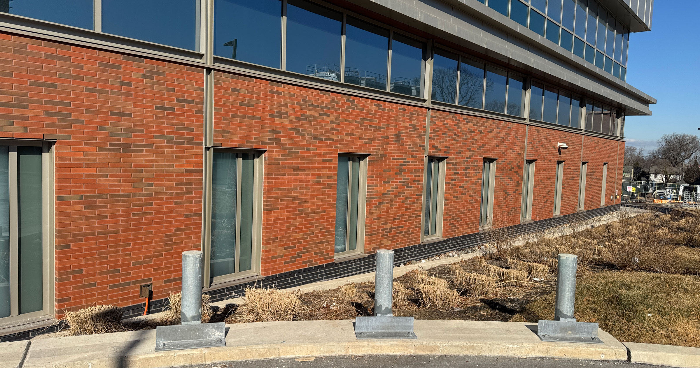 A red brick building with tall, narrow windows, surrounded by brown winter landscaping and four short metal bollards along a curved curb in the foreground. The sky is clear and blue.