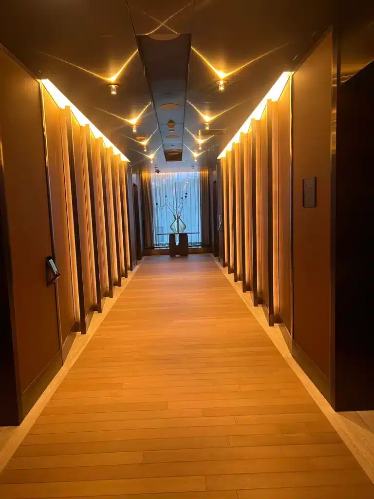 A modern hallway with wooden floors and warm lighting, lined with vertical panels on both sides. At the end, a window with sheer curtains lets in daylight, and a tall vase with flowers sits in front of it.