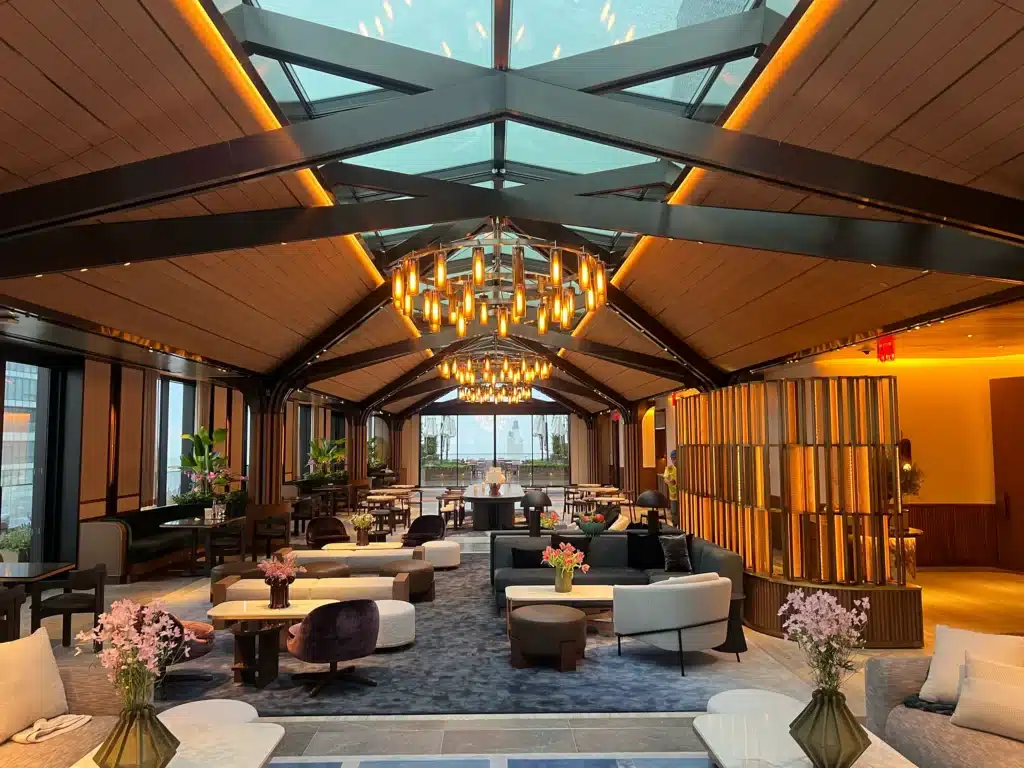 A modern, elegant lounge with a glass ceiling, warm lighting, chandeliers, plush seating, flowers on tables, and large windows letting in natural light. The space has a cozy yet sophisticated atmosphere.