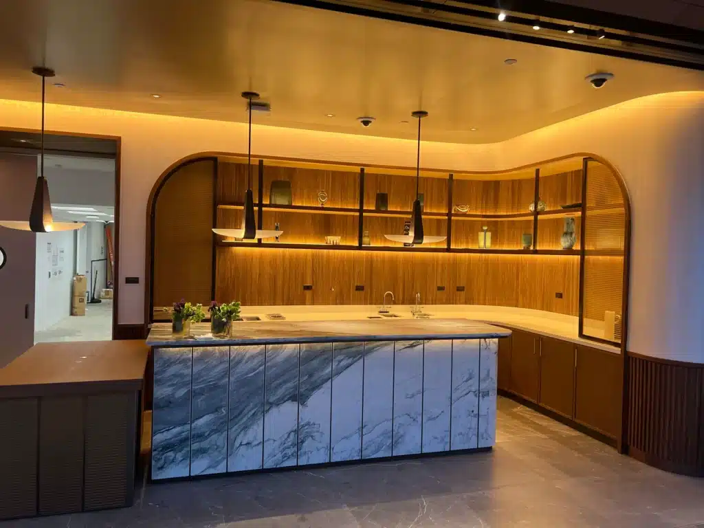 A modern kitchen with warm lighting, a marble island, wooden cabinets, open shelves displaying decor, a sink, and hanging pendant lights. The floor is dark, and a hallway is visible to the left.