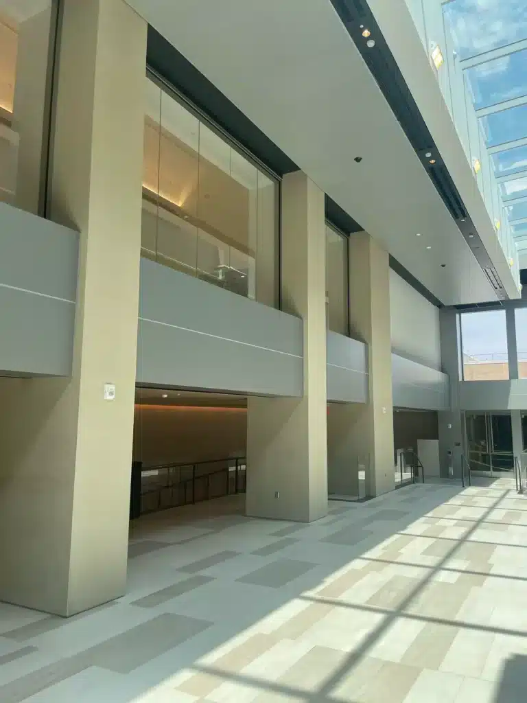 A spacious, modern building interior with high ceilings, large windows, and natural light streaming in. Beige walls, glass railings, and light-colored tiled floors create a clean, open atmosphere.