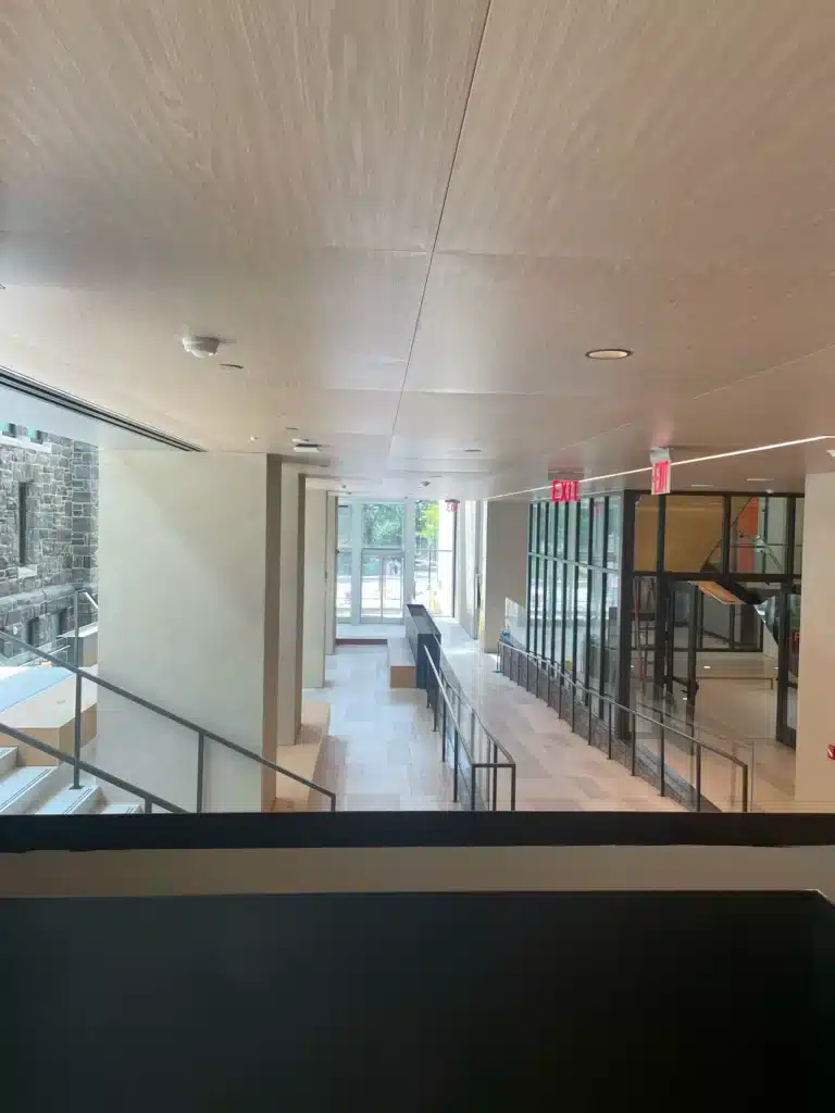 A modern indoor hallway with large windows, light wood ceiling, tiled floors, glass railings, and an exit door visible at the end. Stairs are on the left, and natural light fills the space.