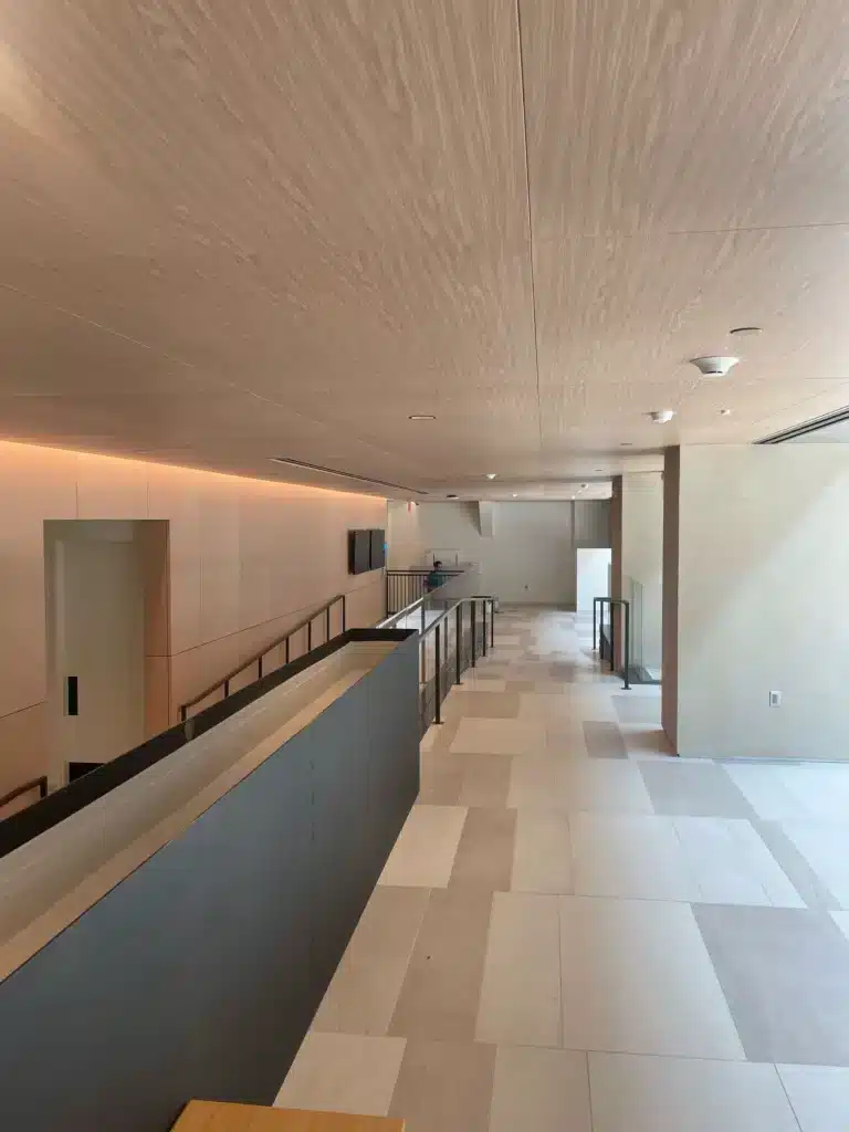 A modern, minimalistic interior with light wood ceilings, beige tile floors, and a long hallway with a black railing. The space is bright and open, with natural light coming through windows at the far end.