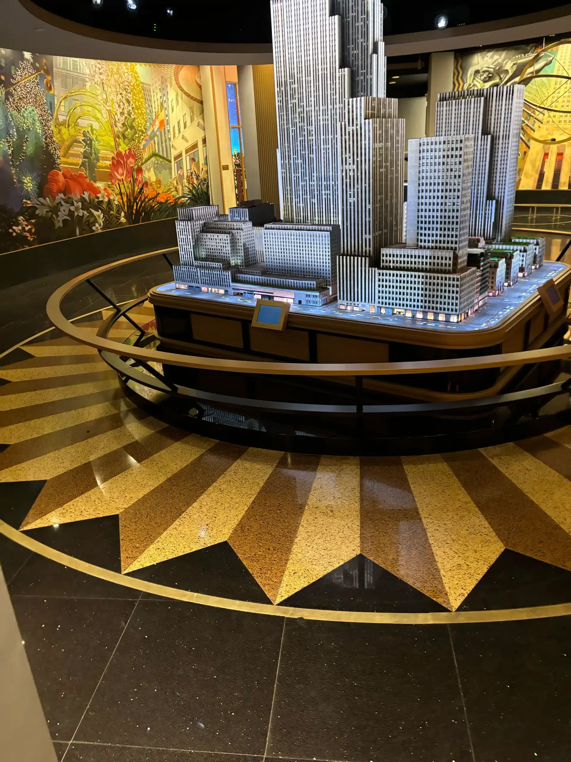 A detailed scale model of the Empire State Building is displayed in a museum, surrounded by a golden star-patterned floor and colorful mural walls.