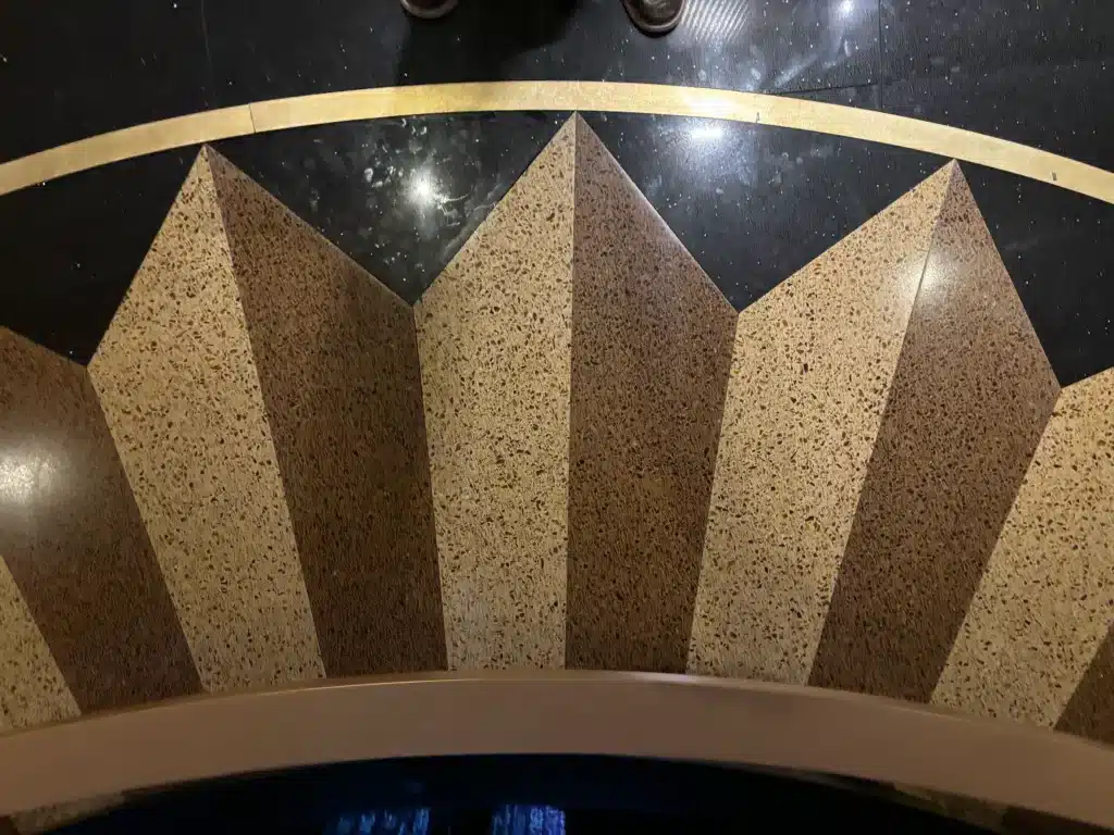 A geometric floor design with alternating light and dark brown stone triangular segments, bordered by a gold stripe on polished black flooring; the tips of brown shoes are visible at the top edge.