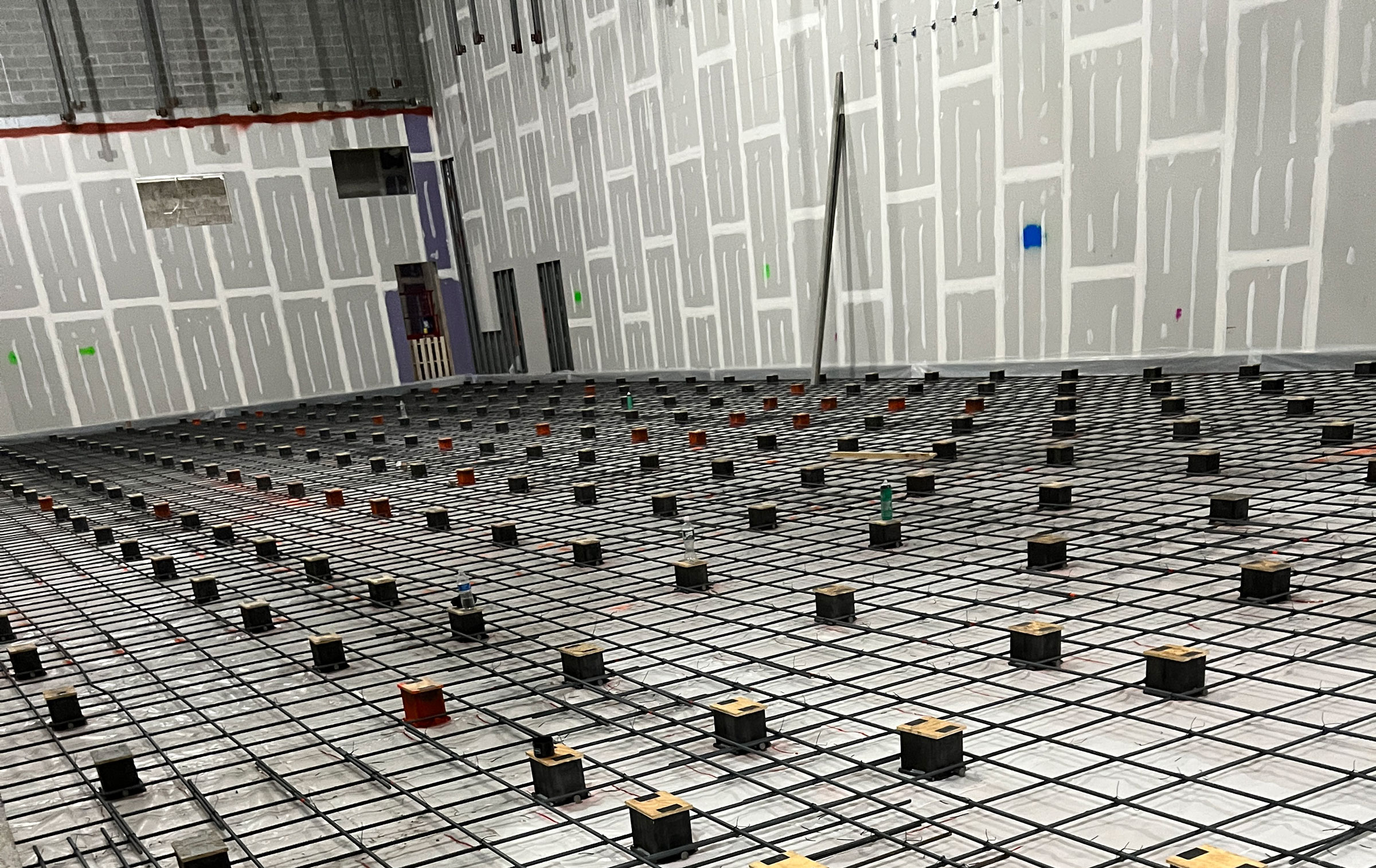 Large indoor construction site with unfinished drywall, metal wall supports, and a grid of rebar on the floor. Numerous small, box-like spacers are evenly placed on the rebar, preparing for a concrete pour.