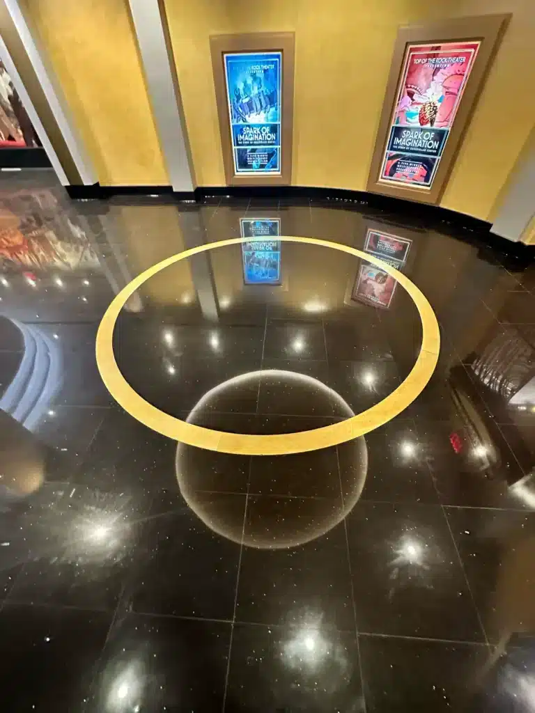 A shiny black floor with a golden ring in the center reflects movie posters and lights from the wall, creating a bright circular reflection within the ring.