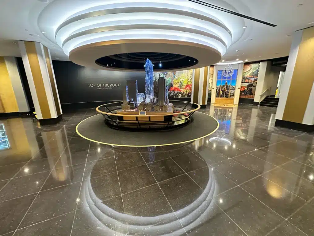 A modern, circular lobby with shiny black floors, a central model of skyscrapers, and a glowing ceiling feature. A wall in the background reads 