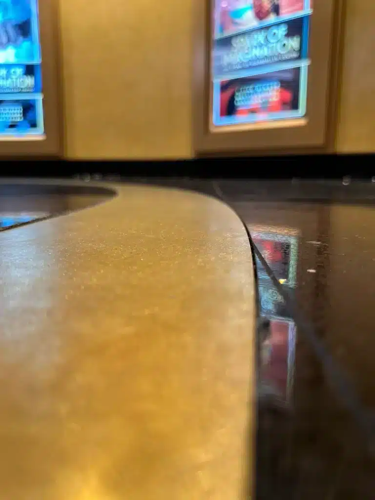 Close-up of a shiny curved counter or table surface in a dimly lit space, with blurred movie posters or digital displays on a gold-colored wall in the background.