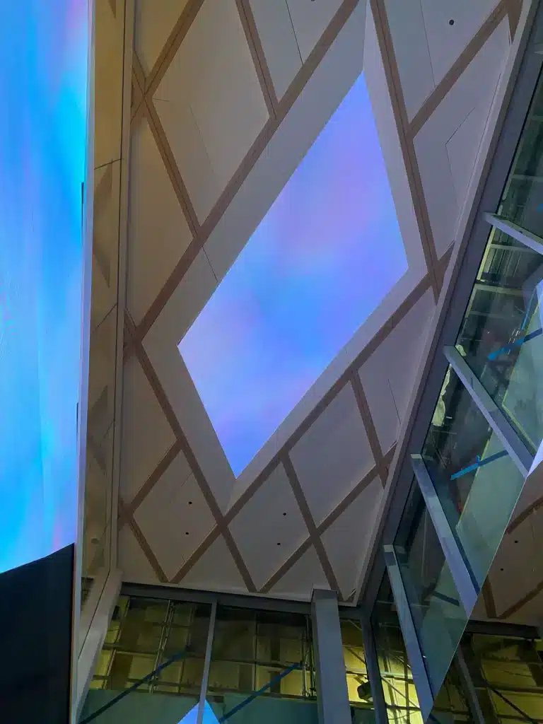 A view looking up at a modern building ceiling with a large rectangular skylight glowing with soft blue and purple hues, surrounded by geometric patterns and glass walls.