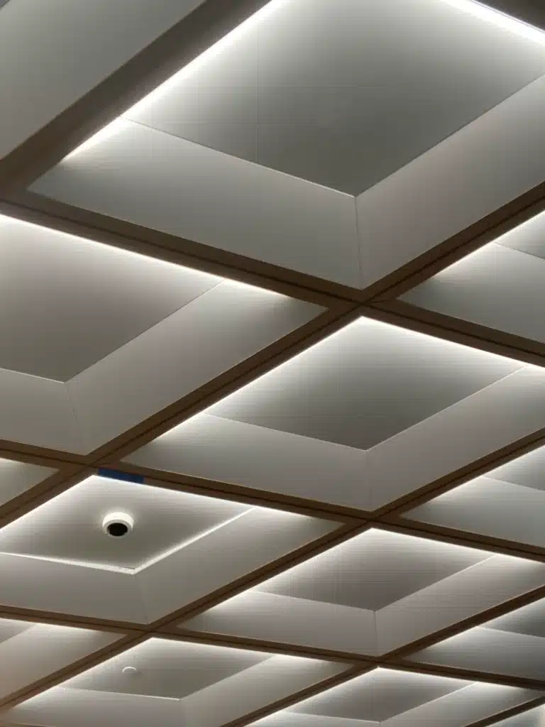 Ceiling with a grid of recessed square panels, each outlined by wood trim and soft white lighting, creating a geometric, modern look. A single security camera is visible between the panels.