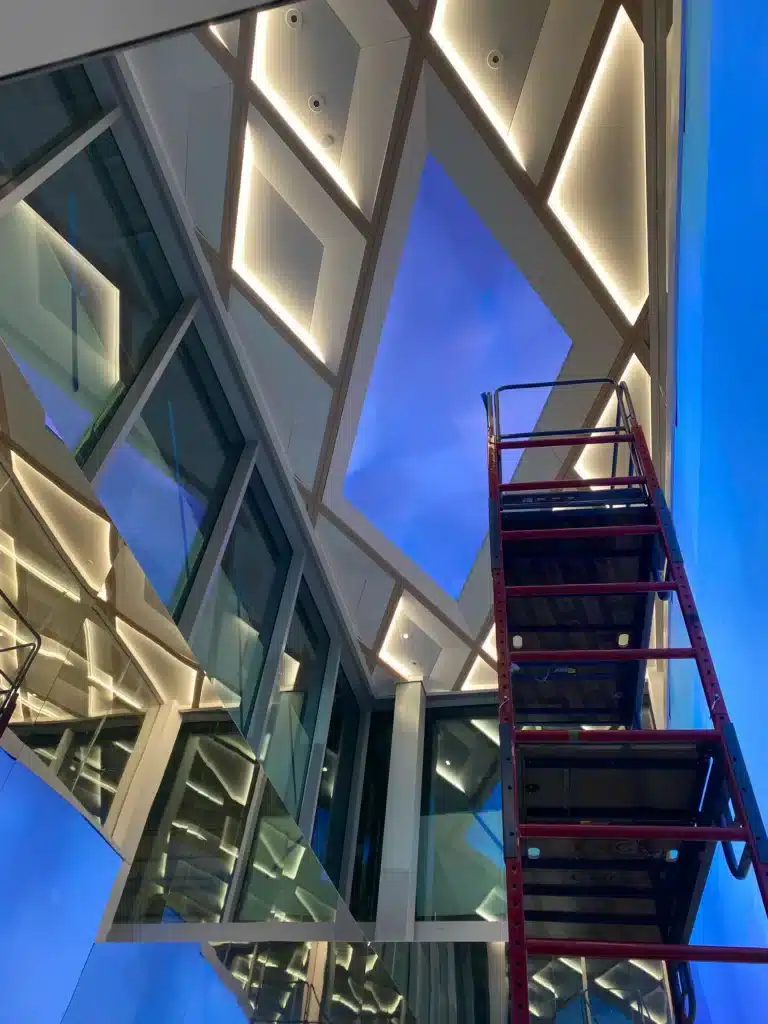 A geometric ceiling with glowing LED lights and glass walls is shown from below. A red metal scaffolding is positioned to the right, and a bright blue sky is visible through an opening in the ceiling.