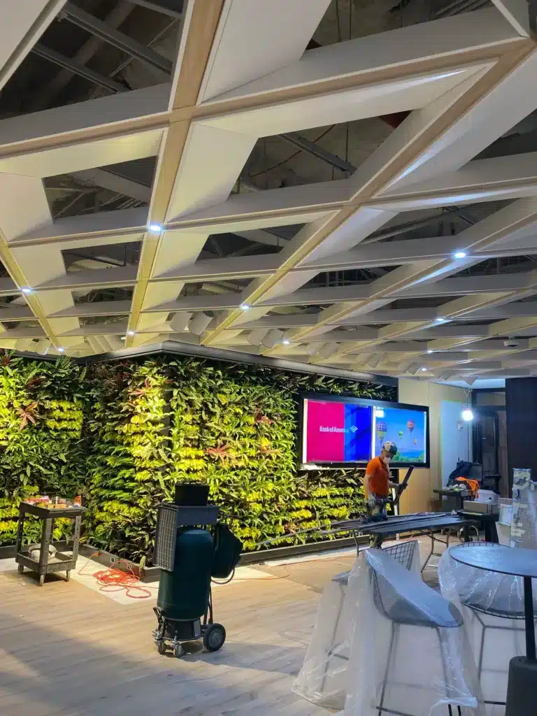 A modern office space under construction features a large vertical green wall, geometric ceiling panels, and workers installing equipment near a screen displaying a colorful welcome message. Tools and furniture are scattered around.