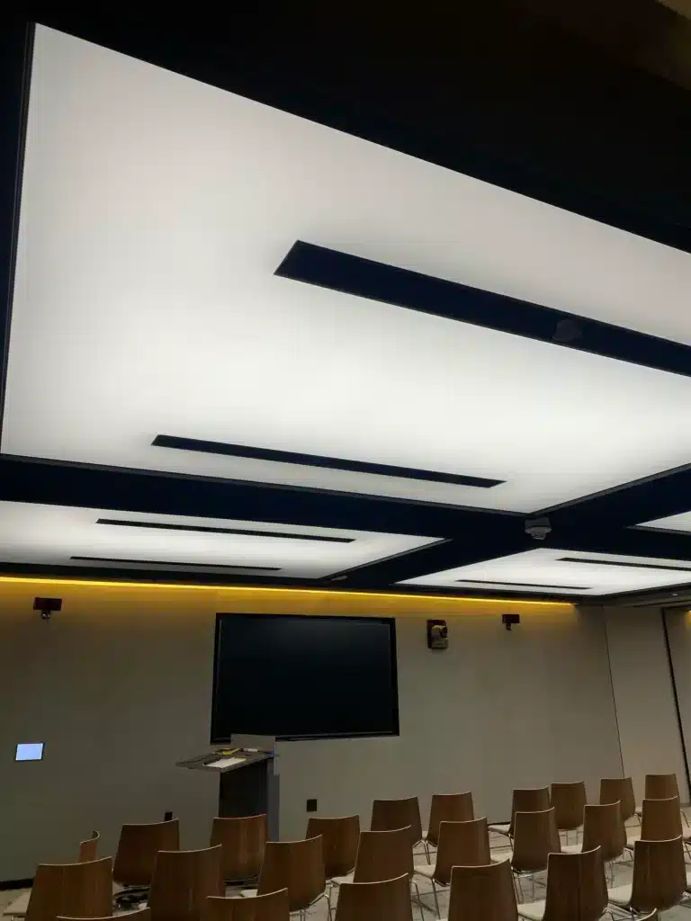 A modern conference room with wooden chairs, a large wall-mounted screen, a podium, and a ceiling featuring geometric, illuminated panels with black accents. The room has a minimalist design and soft lighting.