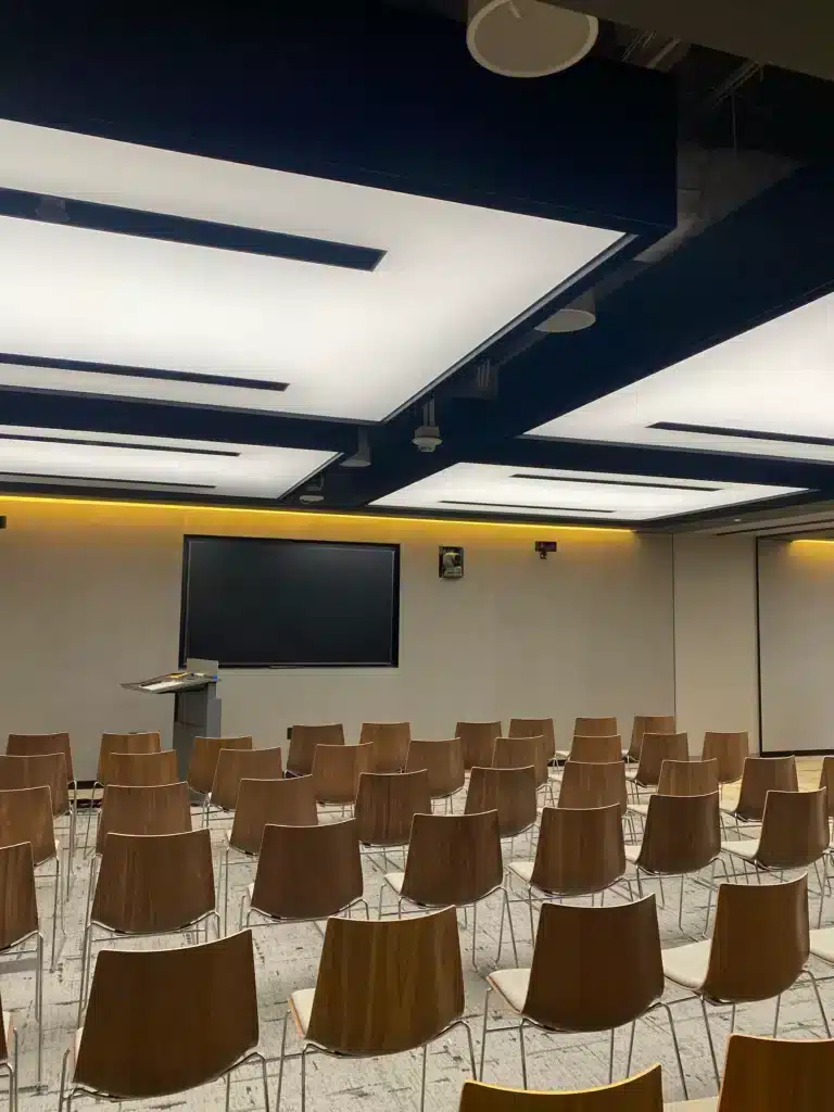 A modern conference room with rows of empty wooden chairs facing a podium and a large blank screen mounted on the wall. The ceiling has large, illuminated panels.