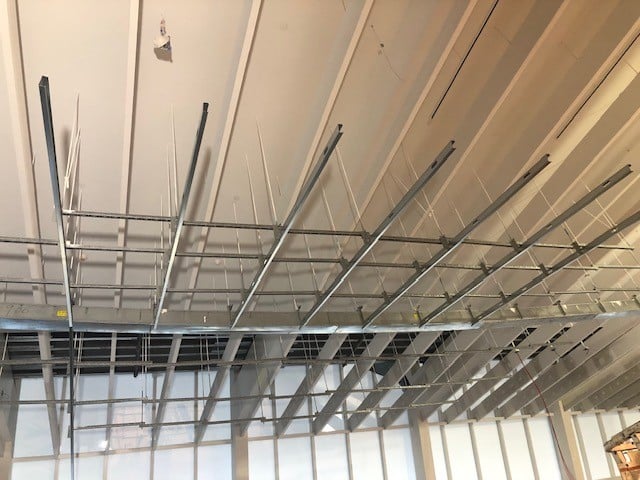 Metal framing and suspension wires for a dropped ceiling installation, with unfinished grid structure and vertical rods, set against a background of large windows and an exposed ceiling.