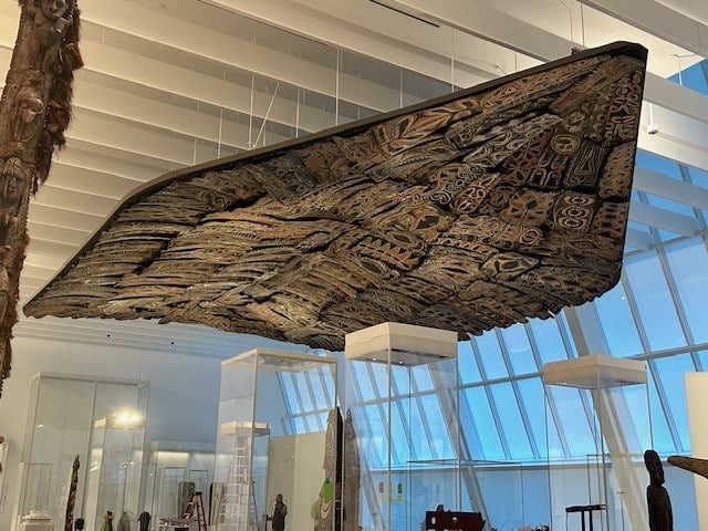 A large, ornately carved wooden canoe is suspended upside down from the ceiling in a bright, modern museum space with tall glass windows and display cases below.