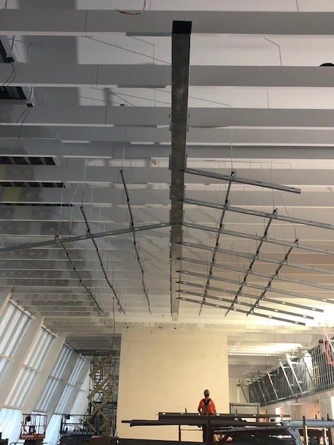 A construction worker stands under a high ceiling with exposed beams and suspended metal framework. Scaffolding and equipment are visible, and large windows line the left wall, letting in natural light.