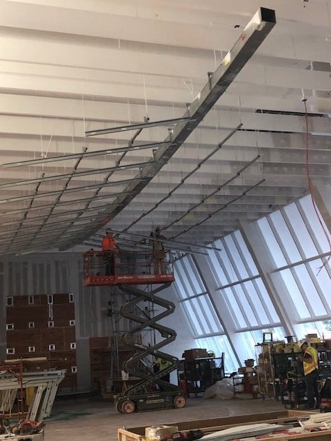 Two construction workers on a scissor lift install metal framework on the ceiling of a large, unfinished building with slanted windows and construction materials scattered around.