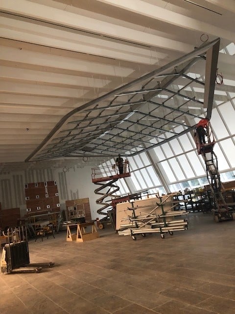 Two workers on scissor lifts install or work on a large, suspended metal and glass structure inside a spacious, unfinished building with high ceilings, large windows, and construction materials scattered around.