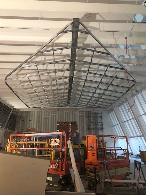 Large indoor construction site with a triangular metal frame structure suspended from the ceiling, scaffolding, construction materials, and orange lifts beneath. White beams and angled windows are visible in the background.