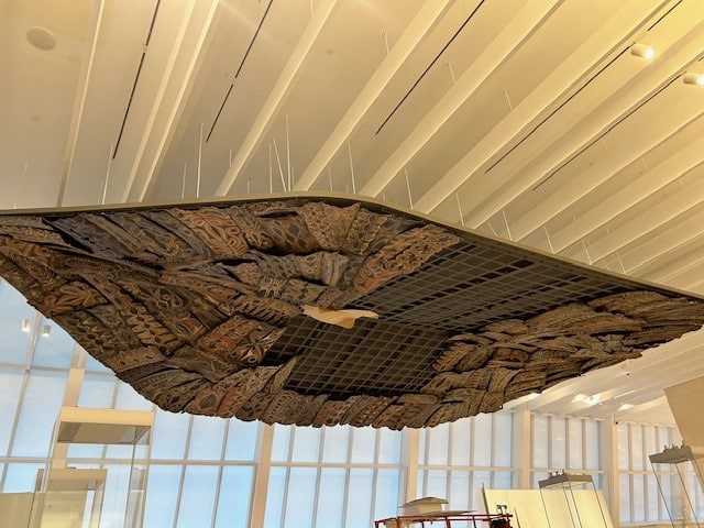 A large, intricately carved wooden panel with geometric and tribal patterns is suspended from the ceiling in a modern, well-lit room with tall windows and white beams.