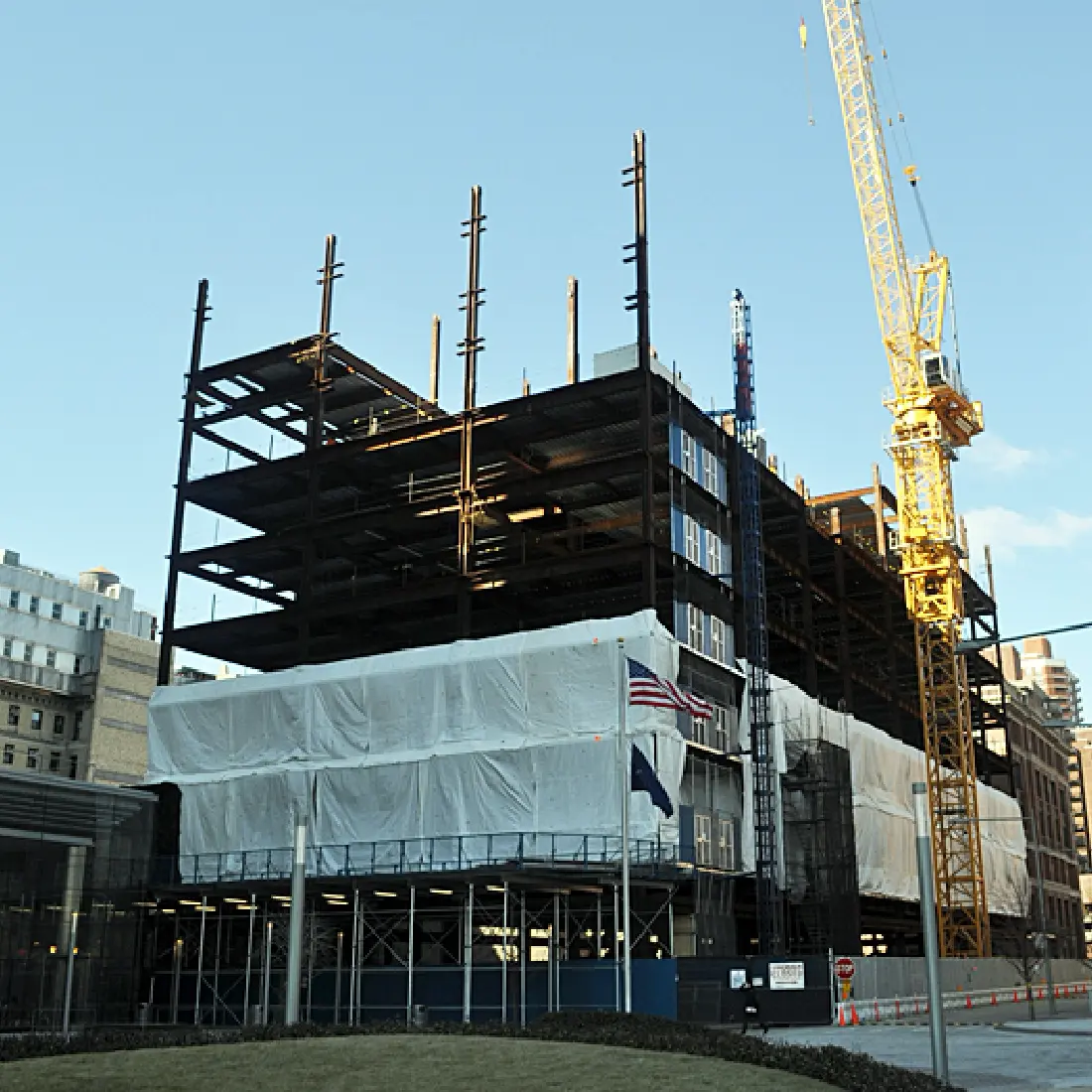 A multi-story building under construction, with exposed steel framework and scaffolding. A large yellow crane stands nearby, and parts of the structure are covered with white sheeting. Two flags are visible in front.