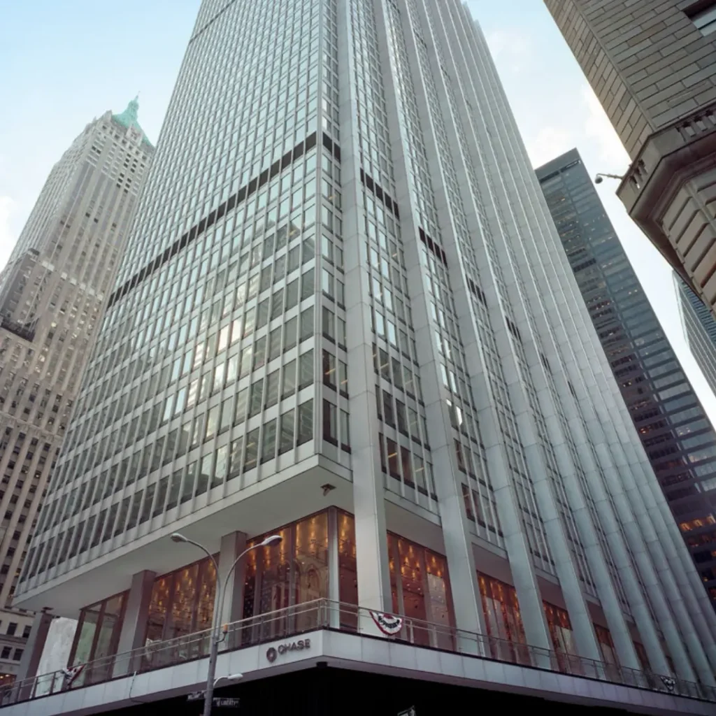 A tall glass and steel office building rises from a city corner, with Chase bank signage visible at its base. The street level is decorated with red, white, and blue bunting. Other skyscrapers surround the building.