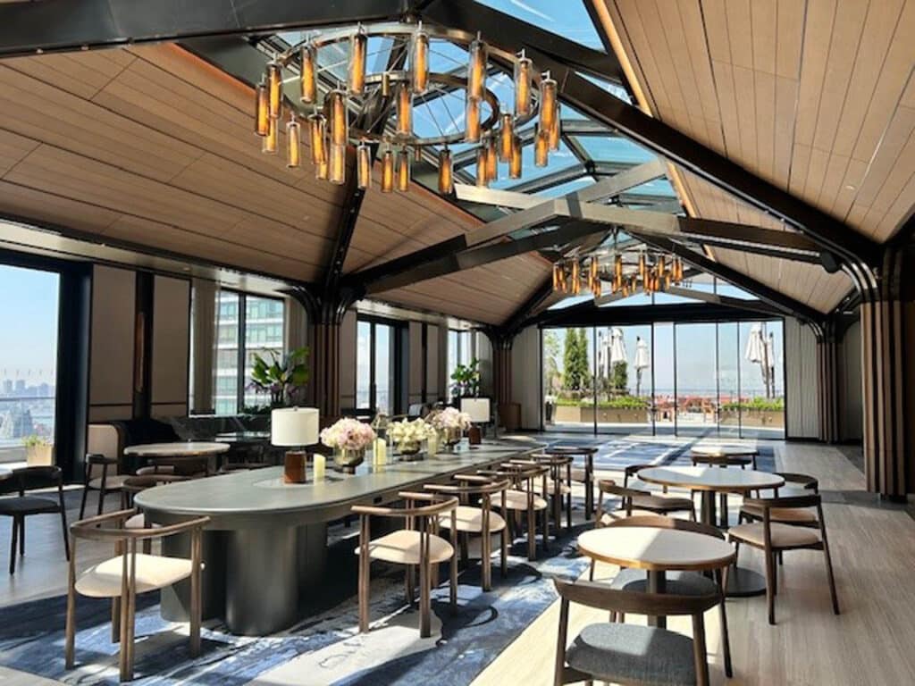 A spacious, modern dining area with large windows, wooden ceilings, elegant chandeliers, and rows of tables and chairs. Sunlight streams in through the glass roof, creating a bright and inviting atmosphere.