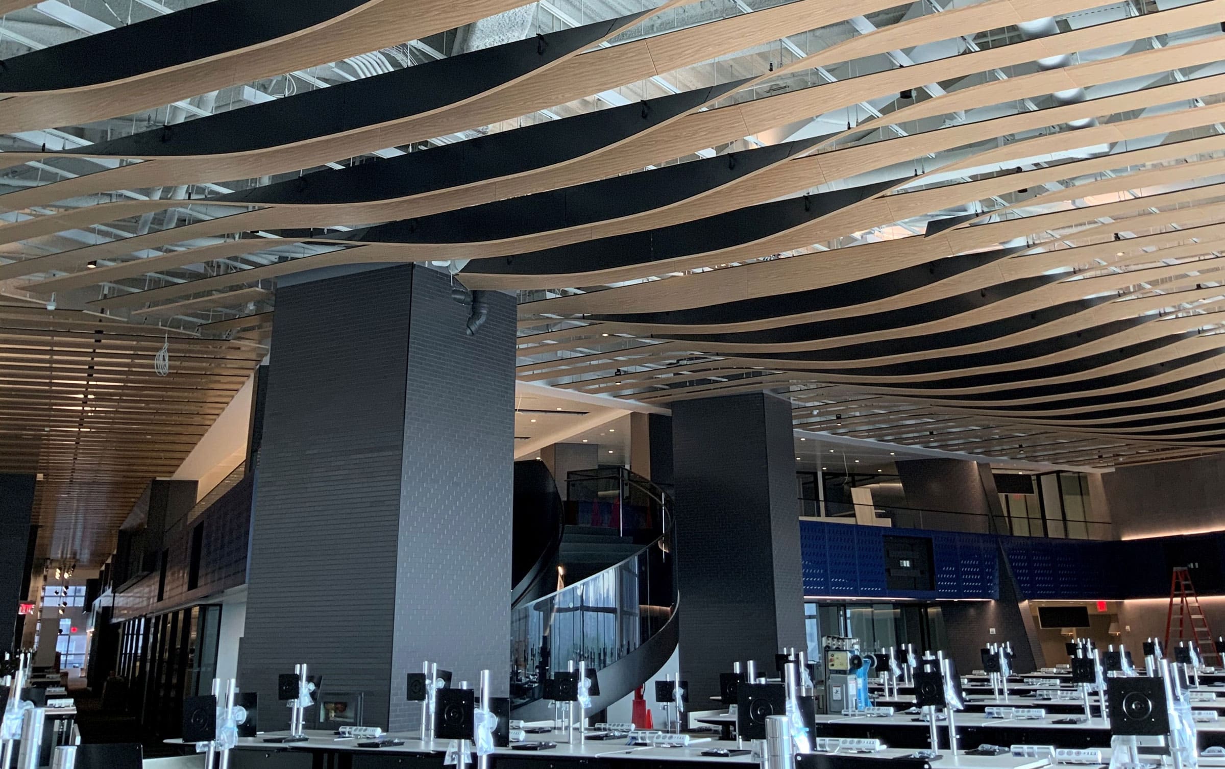Modern interior space with undulating wooden ceiling slats, large pillars, and rows of computer desks. A staircase is visible in the background, and the area appears to be an office or educational setting under construction.