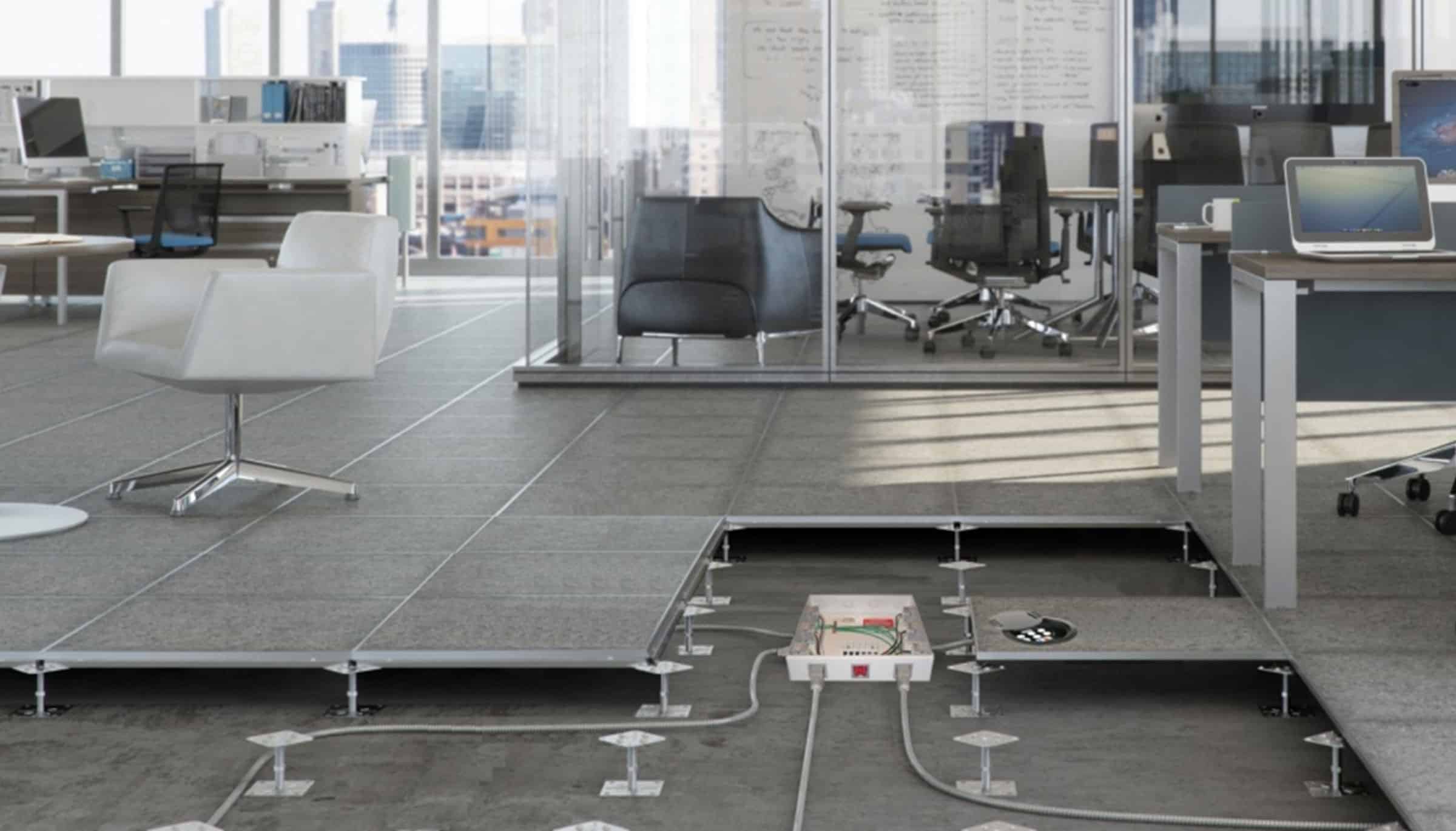 Modern office with desks and chairs, featuring a raised access floor panel removed to show electrical wiring and outlets underneath. Glass-walled meeting room and cityscape visible in the background.